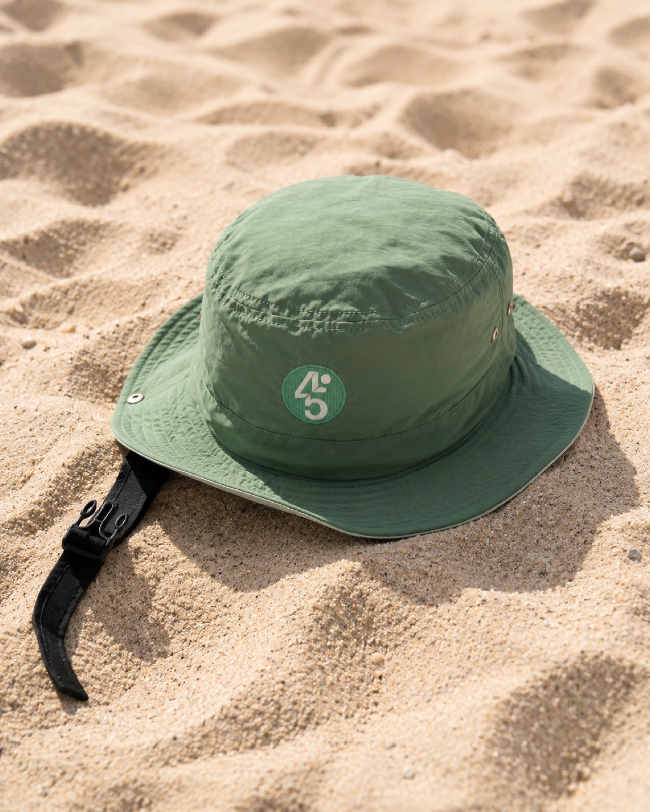 Surf Safe bucket hat resting in the sand, showing the removable helmet insert and chin strap buckle